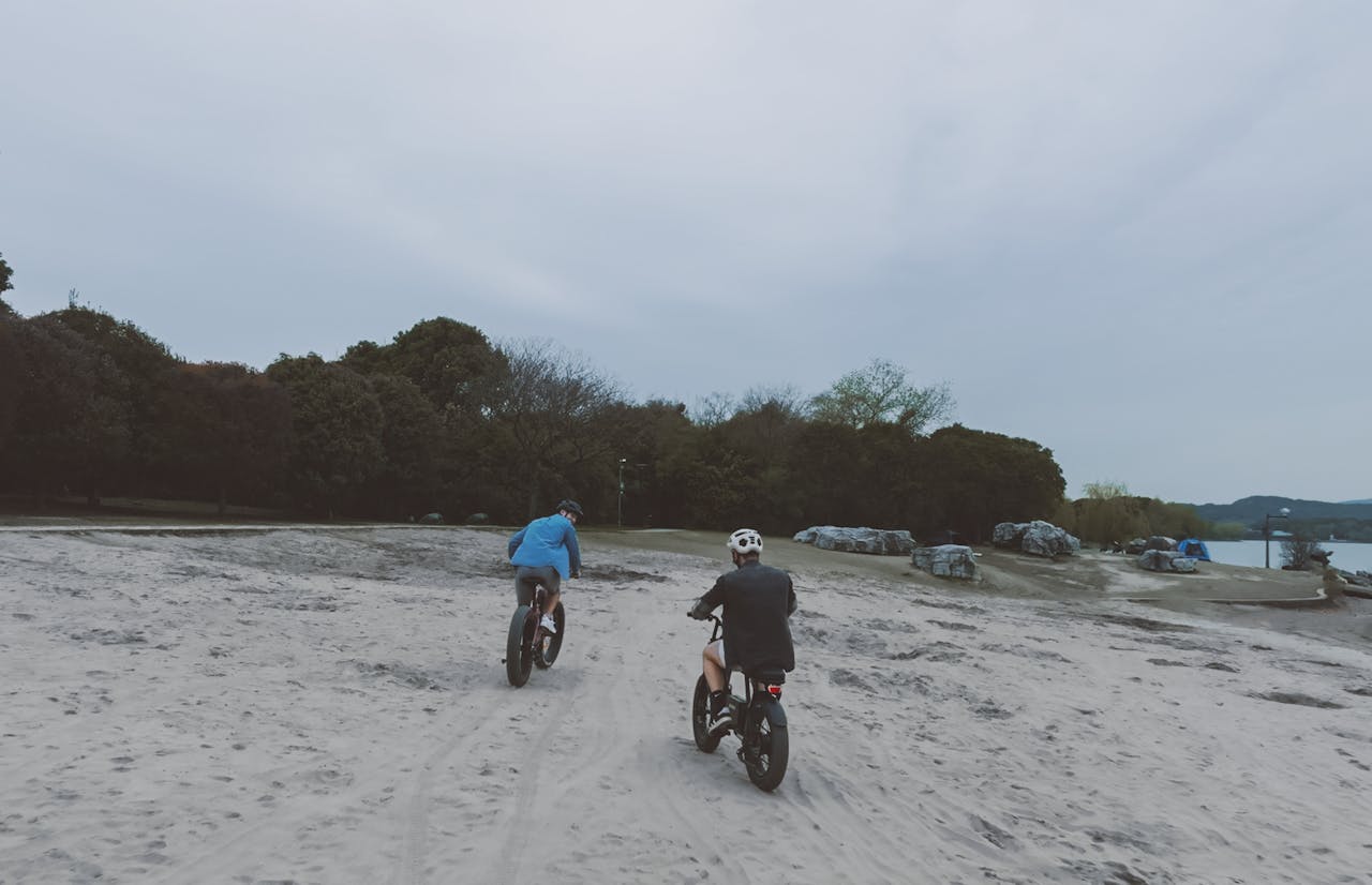 A couple rides bicycles on a sandy beach path on an overcast day, promoting an active lifestyle.